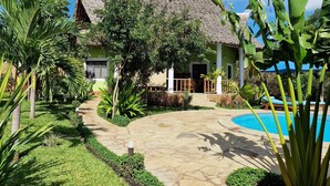 Un resort con un edificio de techo de paja, piscina y un jardín bien cuidado con palmeras y un camino de piedra.