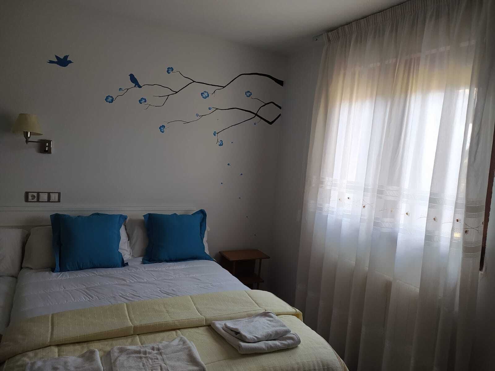 A neatly made bed with blue and white bedding, a wall-mounted lamp, and a branch decoration with birds.