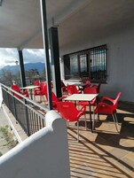 Un espacio al aire libre con sillas rojas y mesas blancas, barandilla y vista a las montañas.
