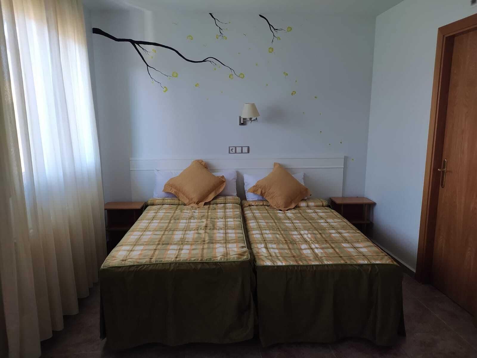 A room with two beds, each with a green bedspread and two pillows. There is a wall-mounted lamp above the beds, and a wooden door on the right.