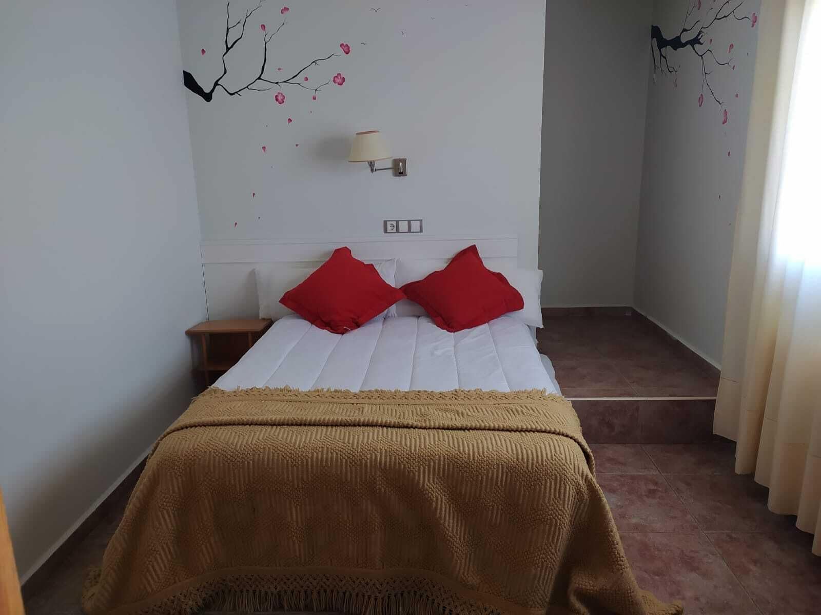 A bedroom with a bed, two red pillows, a beige throw blanket, a nightstand, and a wall-mounted lamp.