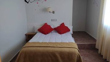 A bedroom with a bed, two red pillows, a beige throw blanket, a nightstand, and a wall-mounted lamp.