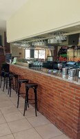 Zona de bar con mostrador de ladrillo, varios taburetes, vasos colgantes y un tablero de menú a la vista.