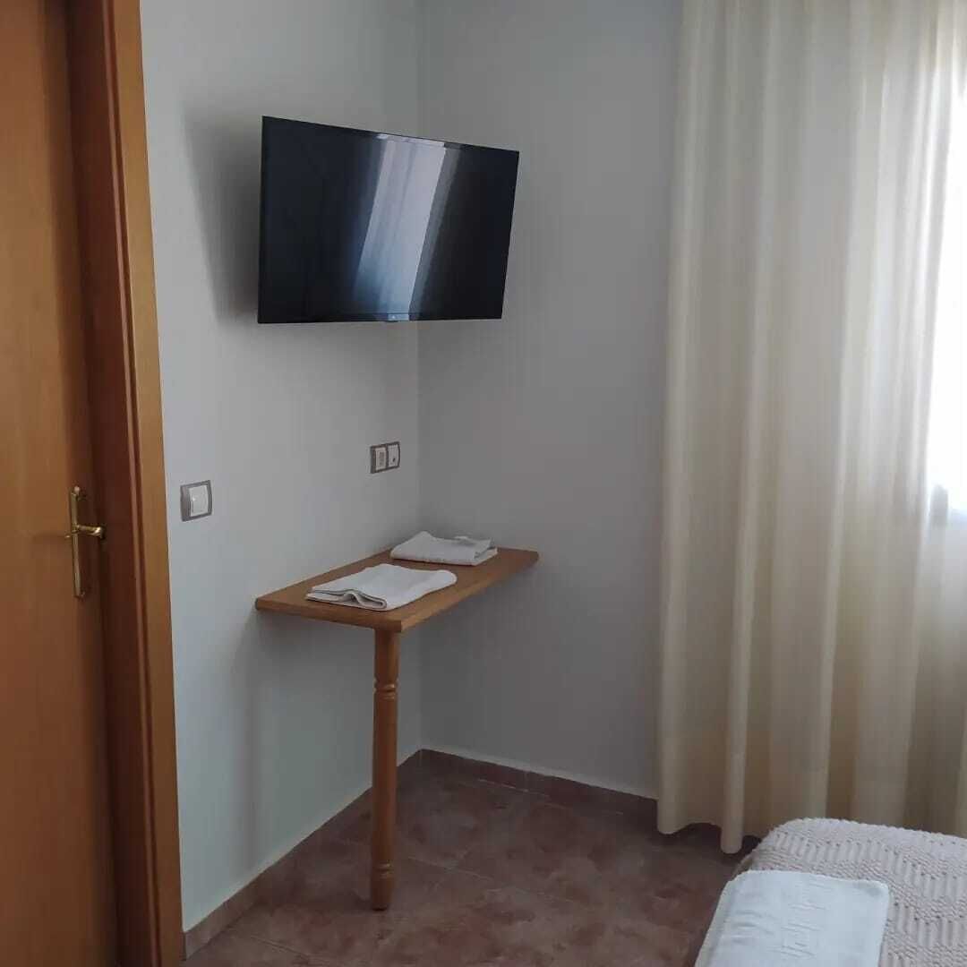 Quarto de hotel com TV de tela plana fixada na parede, mesa de cabeceira de madeira com uma toalha e cama com cobertura branca.