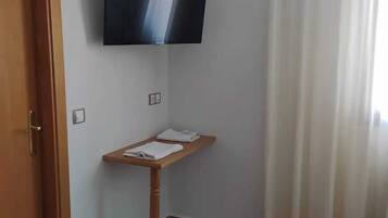 Quarto de hotel com TV de tela plana fixada na parede, mesa de cabeceira de madeira com uma toalha e cama com cobertura branca.