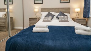 A neatly made bed with a blue cover, two pillows, and a white blanket folded at the foot. There are two bedside tables with lamps, and a large mirror on the left. - 8 Woodlands (Ulverston)