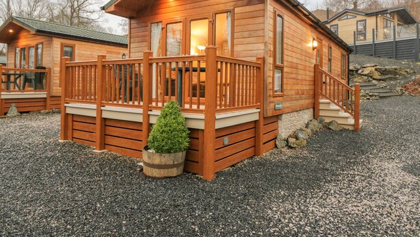 A wooden cabin with a deck, a small potted plant, and a stone pathway. - 8 Woodlands (Ulverston)