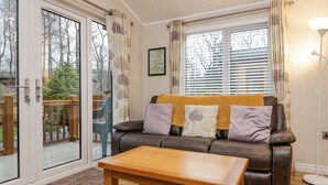 A cozy living room with a leather sofa, wooden coffee table, and a view of a deck and trees through the glass door. - 8 Woodlands (Ulverston)
