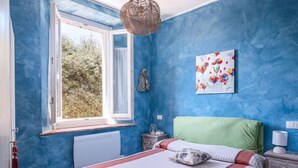 Ein Schlafzimmer mit großem Fenster, einem Bett mit gestreifter Bettwäsche, einem grünen Kopfteil, einem Nachttisch mit Lampe und einem Wandbild.