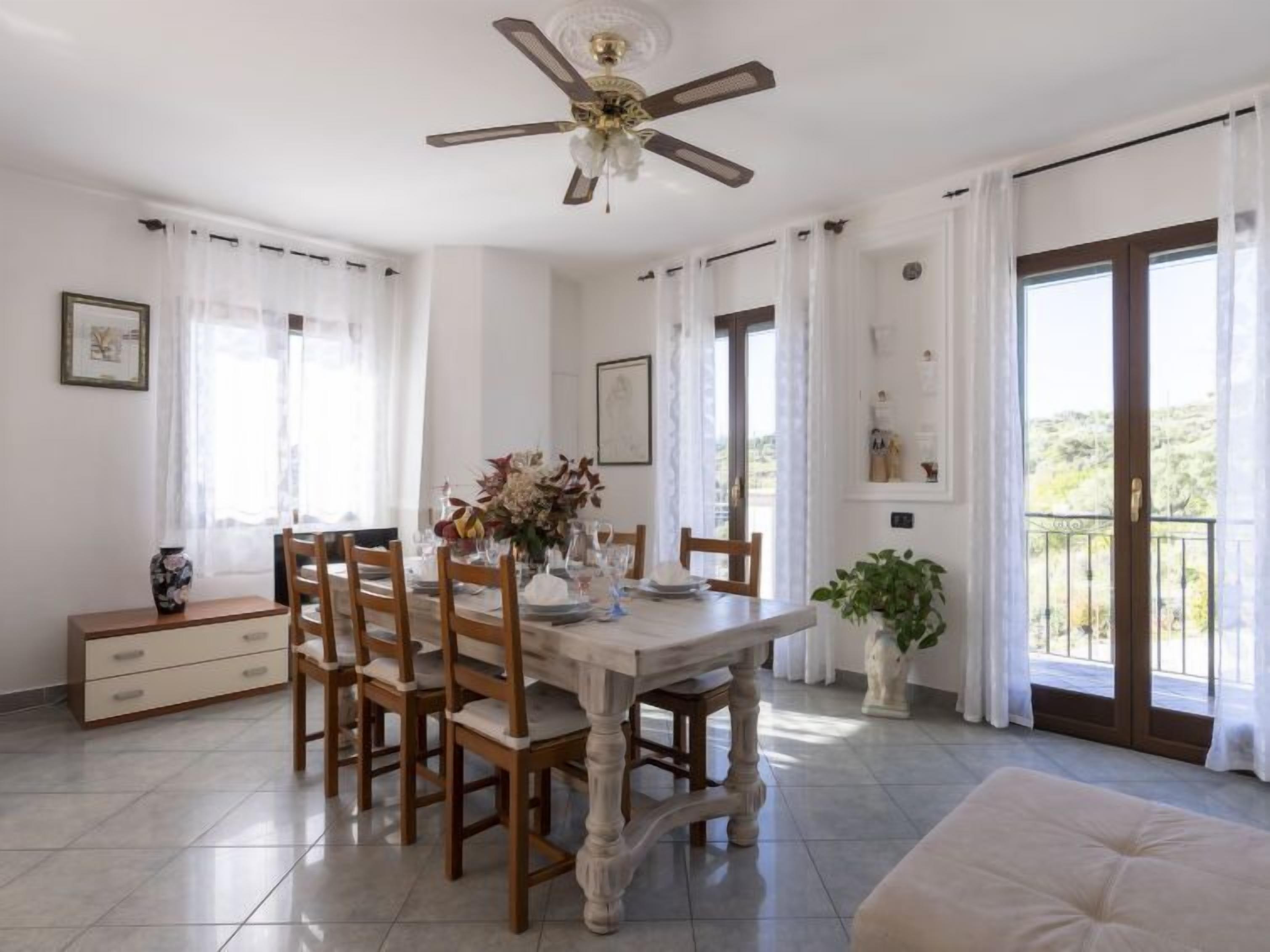 A dining area with a wooden table, chairs, and a ceiling fan. There are windows with curtains, a plant, and a view of the outside.