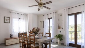 A dining area with a wooden table, chairs, and a ceiling fan. There are windows with curtains, a plant, and a view of the outside. - Apartment Aurora by Interhome (Costarainera)
