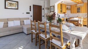 A modern living room with a sofa, dining table, and chairs. There is a kitchen area with wooden cabinets and a ceiling fan. - Apartment Aurora by Interhome (Costarainera)