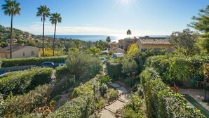 A residential area with a clear view of the ocean, palm trees, and a pathway leading to a building with a tiled roof. - Roche Rouge by Interhome (Théoule sur mer)