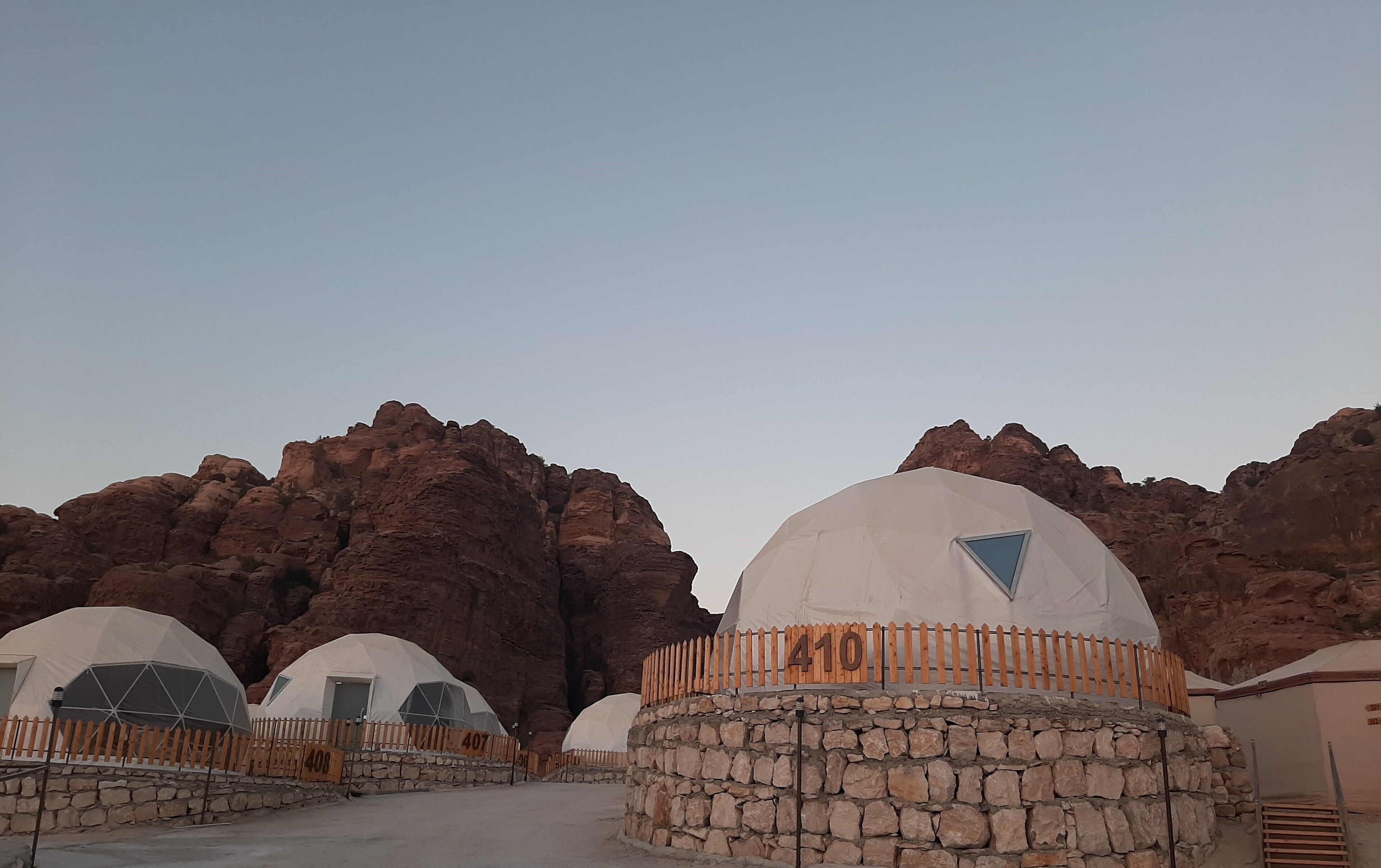 A row of dome-shaped tents with stone walls and wooden fences, set against a backdrop of red rock formations.