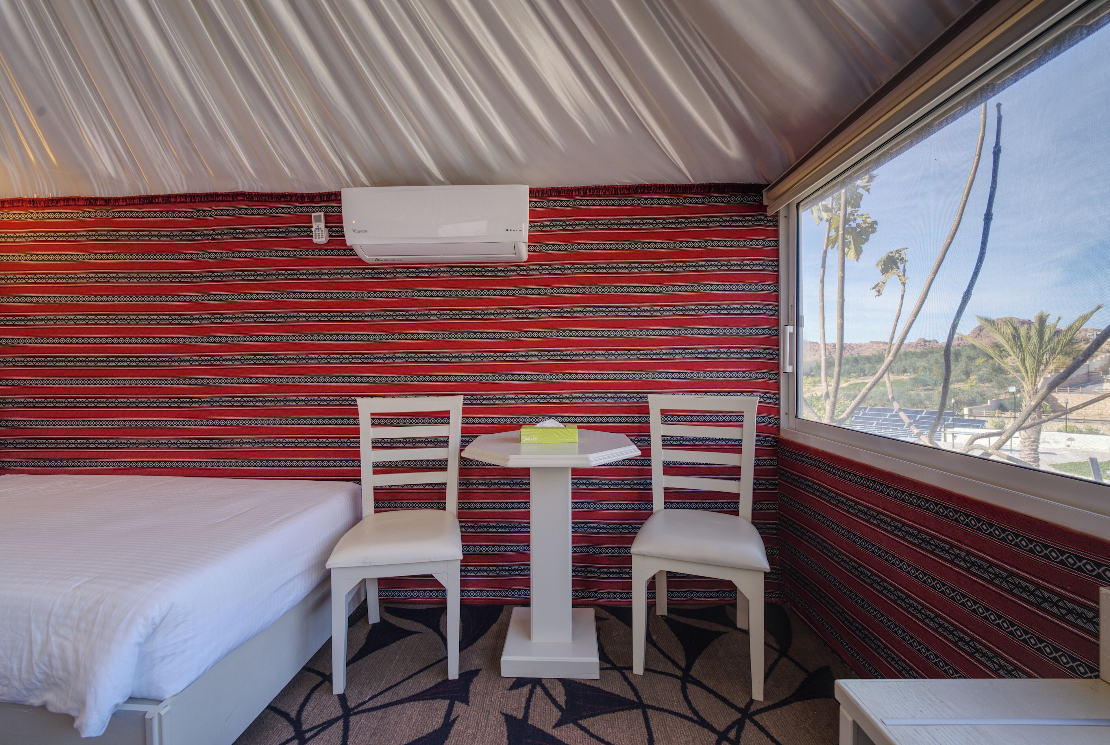 A room with a bed, two white chairs, a small table, and a window with a view of palm trees and mountains.