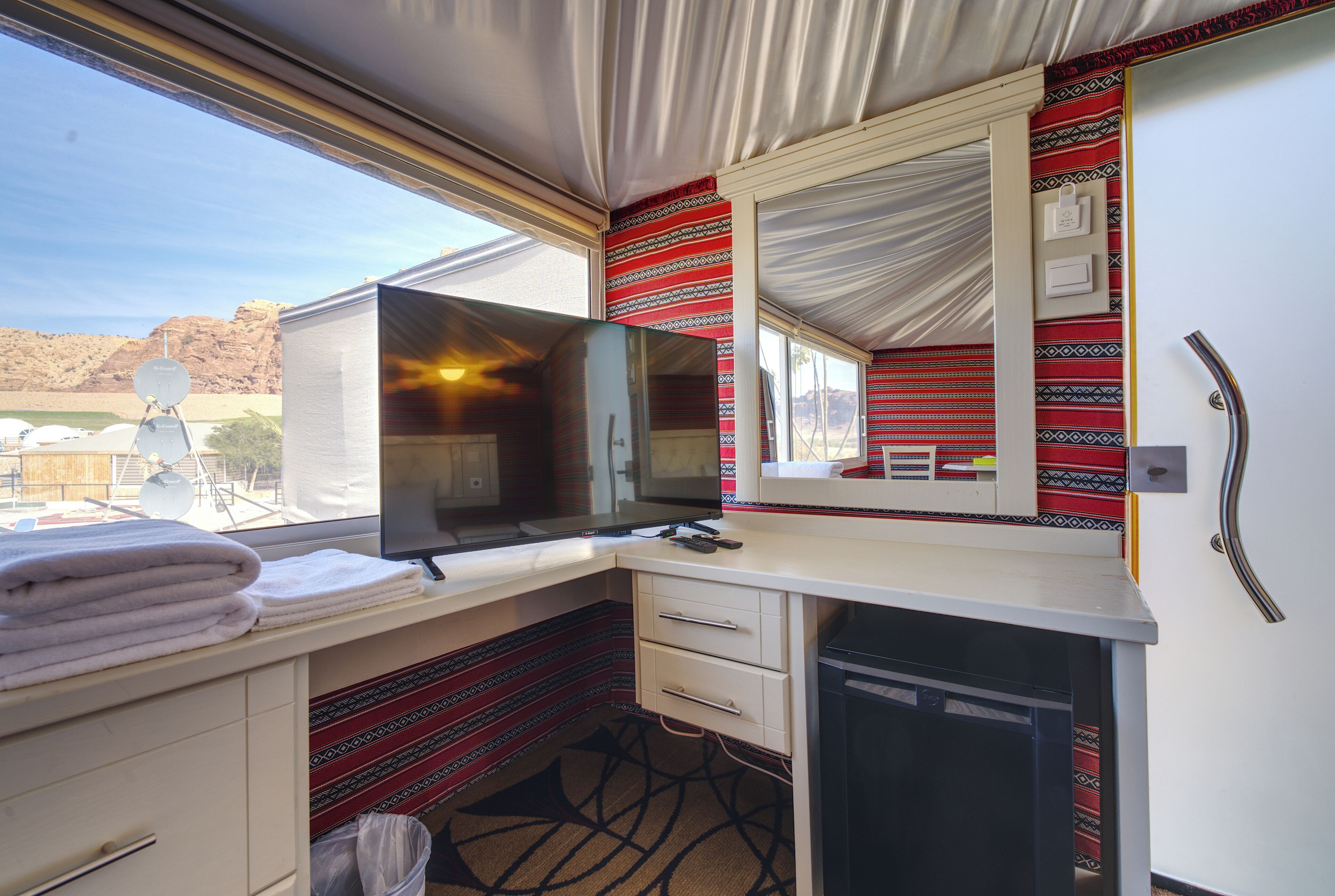 A compact train cabin with a bed, a desk with a computer, a mirror, and a small refrigerator.