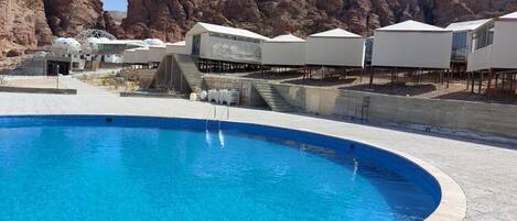 A swimming pool with a curved edge, surrounded by white tents and a rocky landscape in the background.