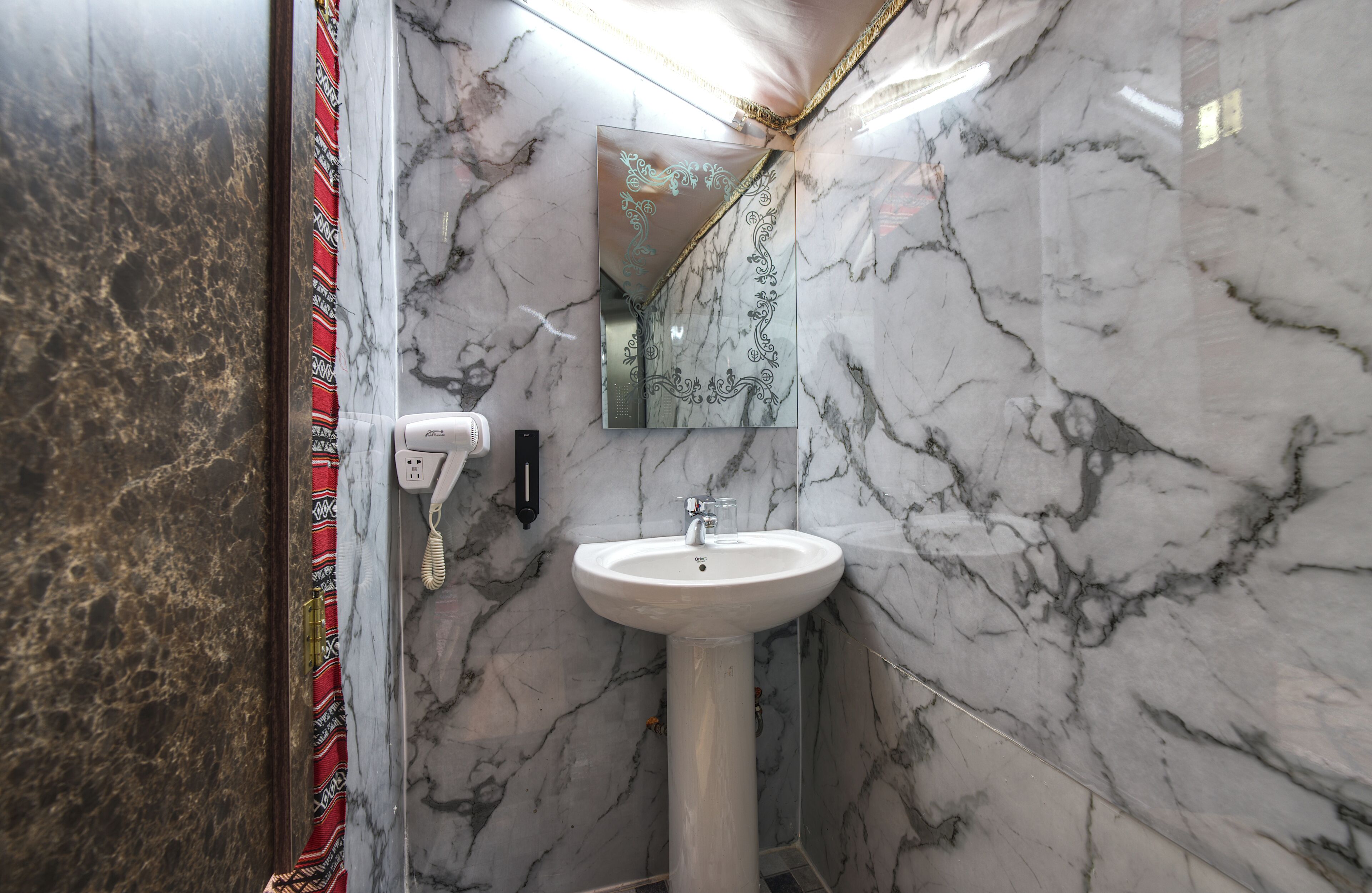 Un baño con lavamanos pedestal blanco, paredes de mármol y espejo con motivos decorativos.
