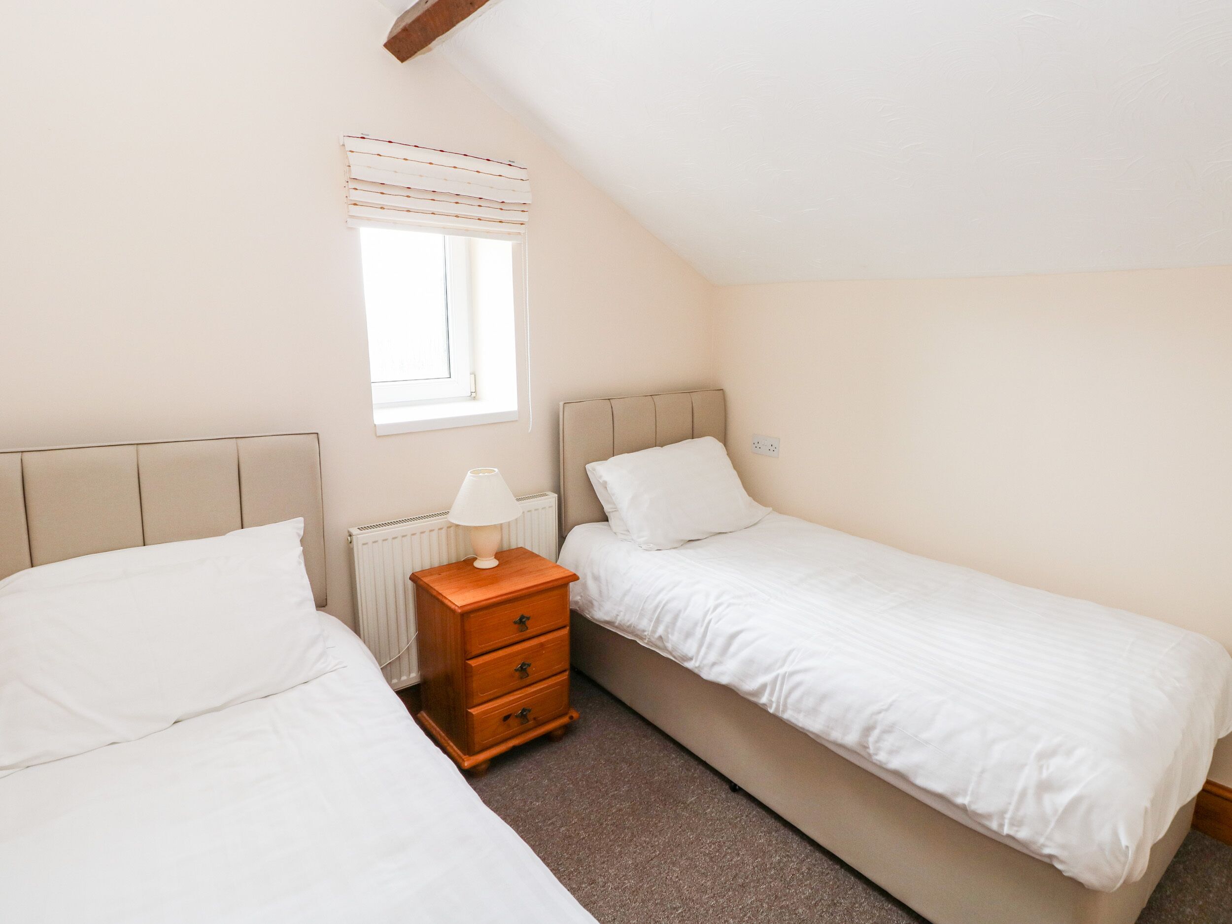 A bedroom with two single beds, a wooden nightstand with a lamp, a radiator, and a window with blinds.