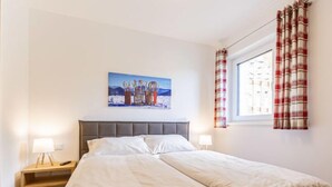 A neatly made bed with white linens, a headboard, and a painting of a winter scene above it.
