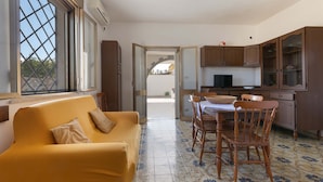 A living room with a yellow sofa, a dining table with chairs, and a kitchen area with wooden cabinets and a TV. - 3231 Villino San Basilio by Barbarhouse (Melendugno)