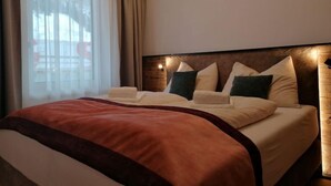 Une chambre d’hôtel avec un grand lit, une literie de couleur beige et un plaid marron. Il y a une fenêtre avec des rideaux légers et une vue sur un bâtiment à l’extérieur.