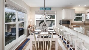 Un comedor con una mesa blanca dispuesta para seis personas, una silla blanca en primer plano, y una zona de cocina con un microondas y un ventanal.