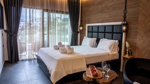 Quarto de hotel com uma cama grande, uma mesinha com uma bandeja de frutas e copos, uma cadeira e vista para o exterior através de janelas amplas.