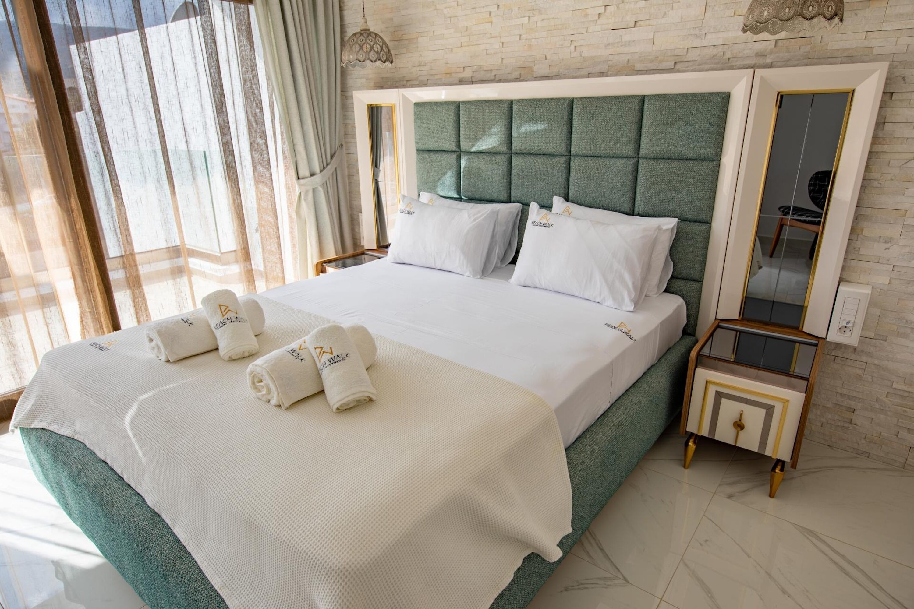 A neatly made bed with white linens and towels, a green upholstered headboard, and a small bedside table with a mirror.