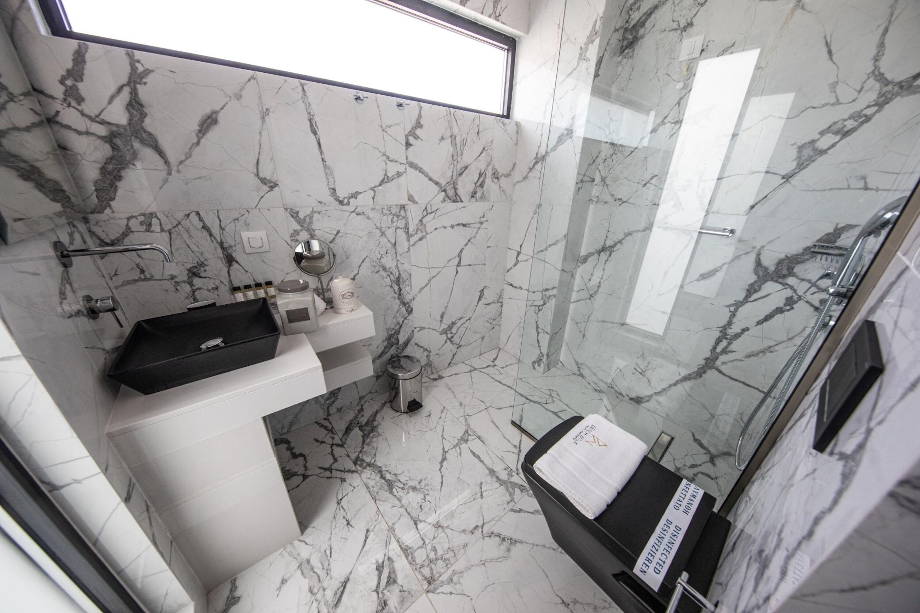 A modern bathroom with marble walls and floor, a glass shower enclosure, a black sink with a soap dish, and a towel rack.
