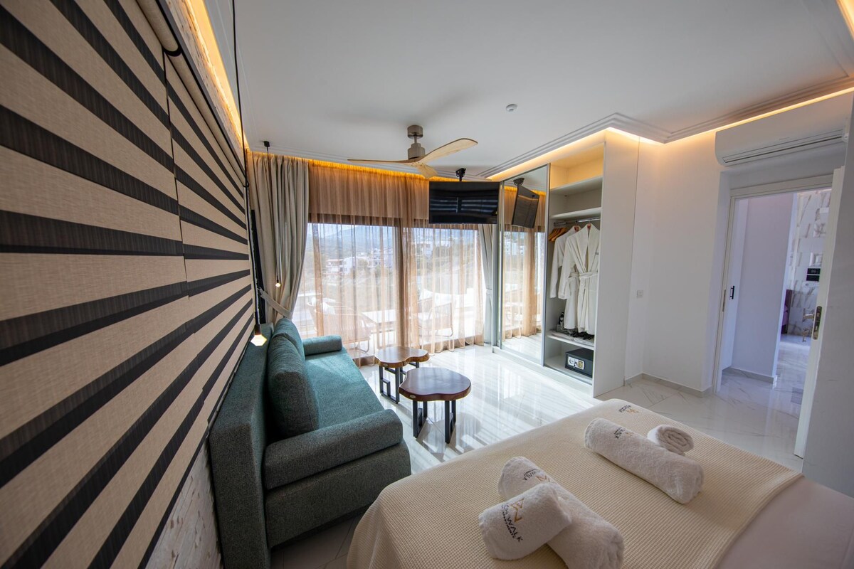 A hotel room with a sofa, a small table, a bed with towels, a ceiling fan, and a view through the window.