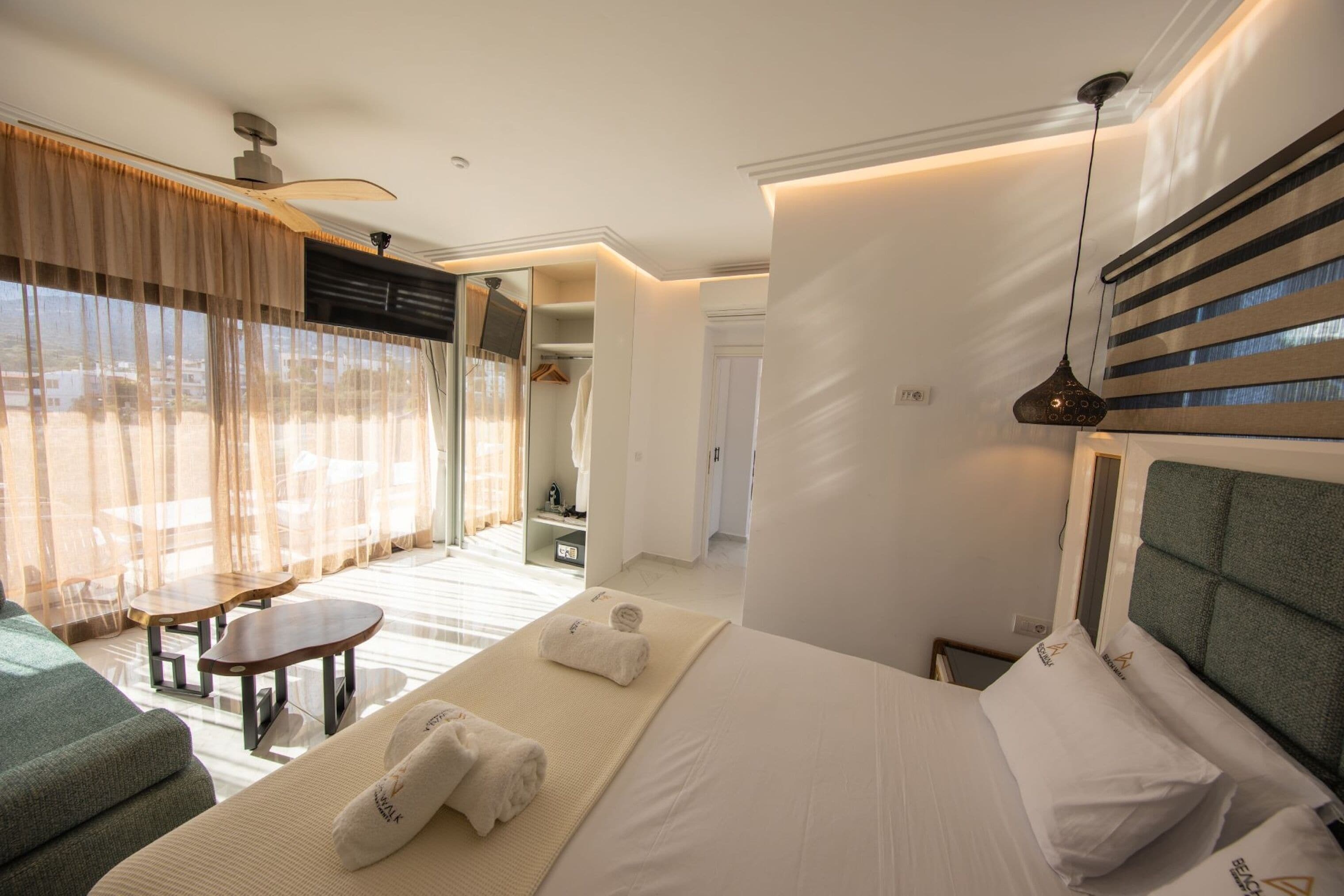 A hotel room with a bed, a sofa, a small table, and a chair. There is a window with curtains, a ceiling fan, and a hanging light fixture.
