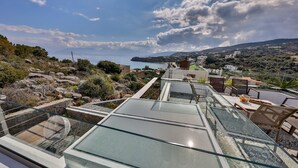 Terraço com guarda-corpo de vidro com vista para a paisagem costeira, incluindo área de estar ao ar livre e vista para o mar.