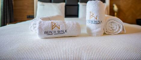 En snyggt bäddad säng med två handdukar, en upprullad och en vikt, och båda försedda med logotypen för ”Beach Walk Luxury Apartments”.
