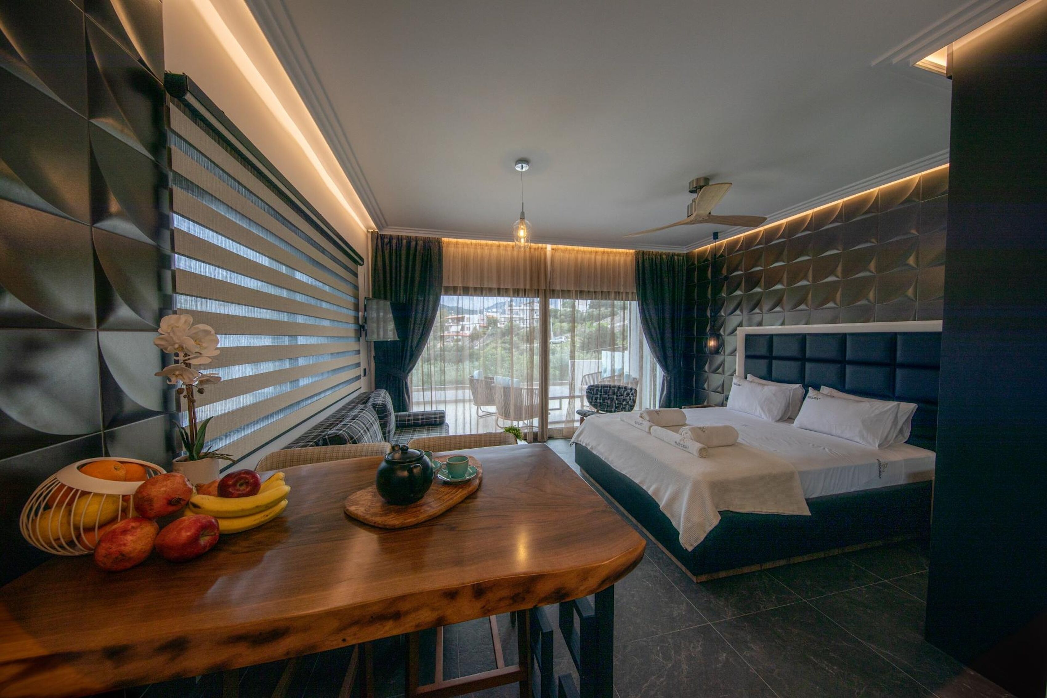 A modern hotel room with a bed, a wooden table with fruits and a teapot, a ceiling fan, and a view of the outdoors.