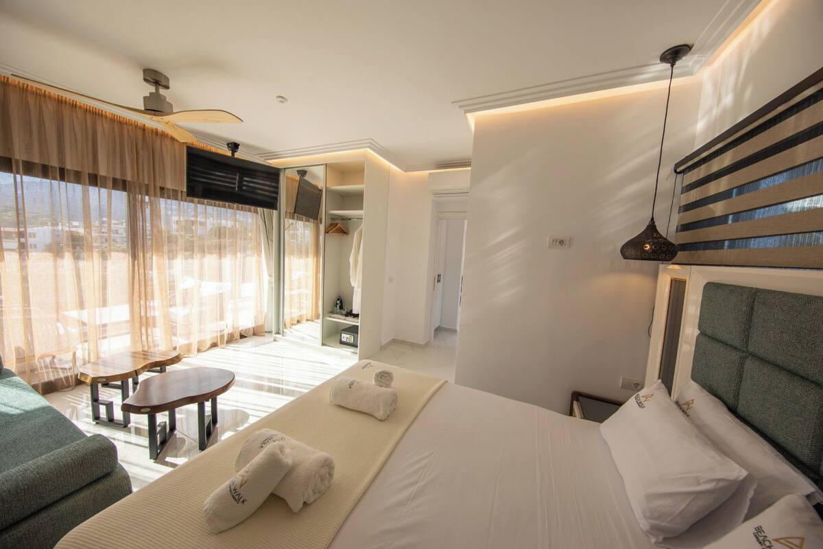 A hotel room with a bed, a small table, a sofa, a window with curtains, and a hallway.