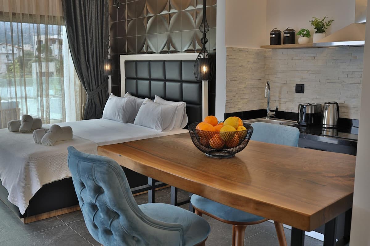 A modern hotel room with a neatly made bed, a wooden dining table with a fruit bowl, and a kitchen area with a coffee maker.