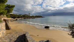 Praia costeira com um muro de pedra e vista para o mar e montanhas ao longe.