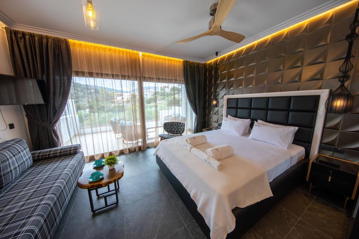 A modern hotel room with a large bed, a sofa, a small table with a plant, and a ceiling fan.