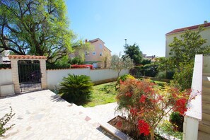 Une cour avec un sol en pierre, une porte en métal et diverses plantes et arbustes.