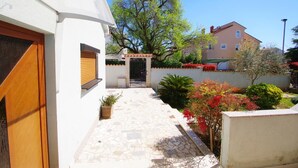 Un étroit sentier mène à une entrée fermée par un portail, qui est elle-même entourée de diverses plantes et d’un grand arbre.