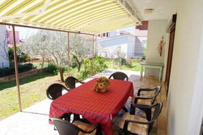 Une terrasse couverte avec une table dressée pour quatre personnes, entourée de chaises, et une vue sur un jardin et des bâtiments en arrière-plan.
