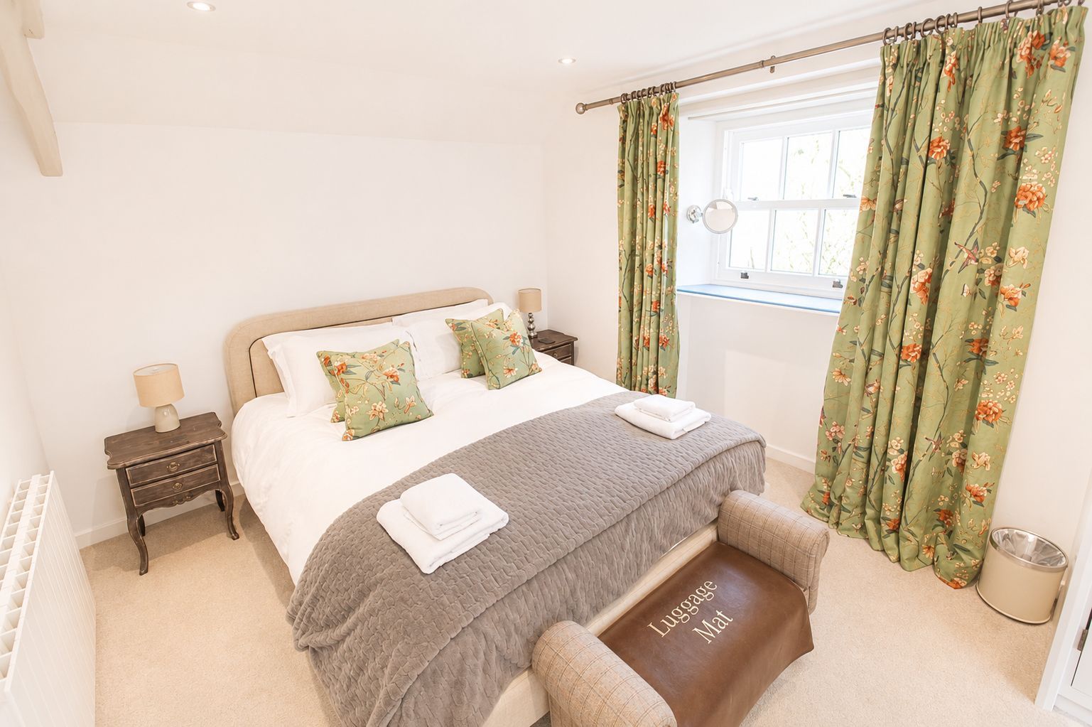A neatly made bed with floral pillows, a bedside table with a lamp, and a window with curtains.