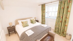 Een netjes opgemaakt bed met bloemenkussens, een nachtkastje met een lamp en een raam met gordijnen.