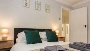 A neatly made bed with white and green pillows, a folded grey blanket, and a bedside lamp.