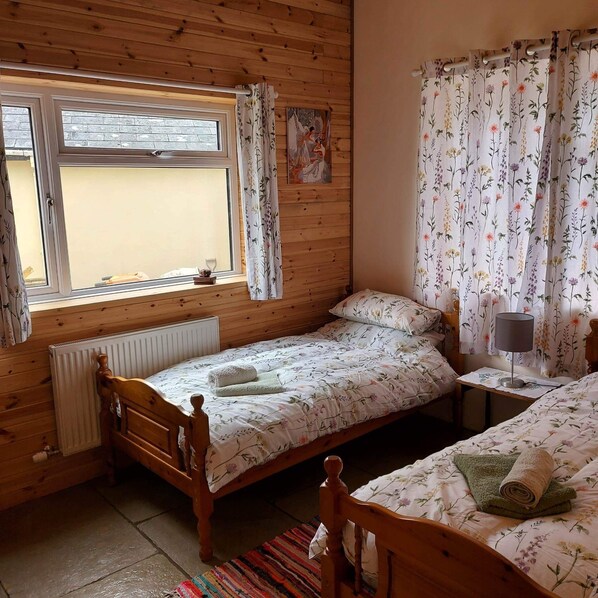A wooden room with two single beds, a window with curtains, a radiator, and a small table with a lamp.
