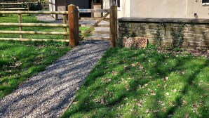 A rustic building with a stone foundation and a green roof, surrounded by a wooden fence and a gravel path leading to a wooden gate. - Sycamore Lodge-1 Bed-sleeps 4-free Parking (Hereford)