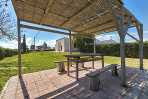 Un espace extérieur abrité, doté d’une table de pique-nique et de bancs, entouré d’une terrasse en briques et d’une pelouse.