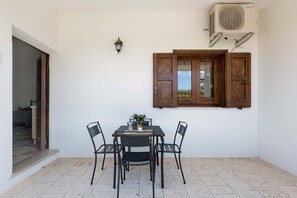 Une terrasse avec une table et des chaises, une fenêtre avec des volets en bois, un appareil de climatisation et un luminaire fixé au mur.