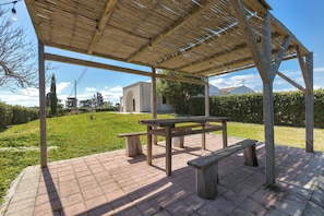 Un espacio al aire libre cubierto con una mesa de picnic y bancos, rodeado por un patio de ladrillo y un césped.